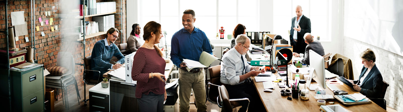 Open-plan office with diverse colleagues working and collaborating at desks; some people are talking, others are using computers or reading documents, in a bright room with large windows and shelves.