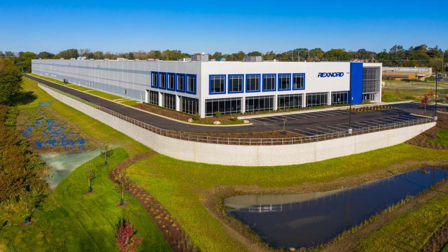 A large, modern industrial building labeled REXNORD with blue accents, surrounded by green lawns, a pond, and an empty parking lot under a clear blue sky.