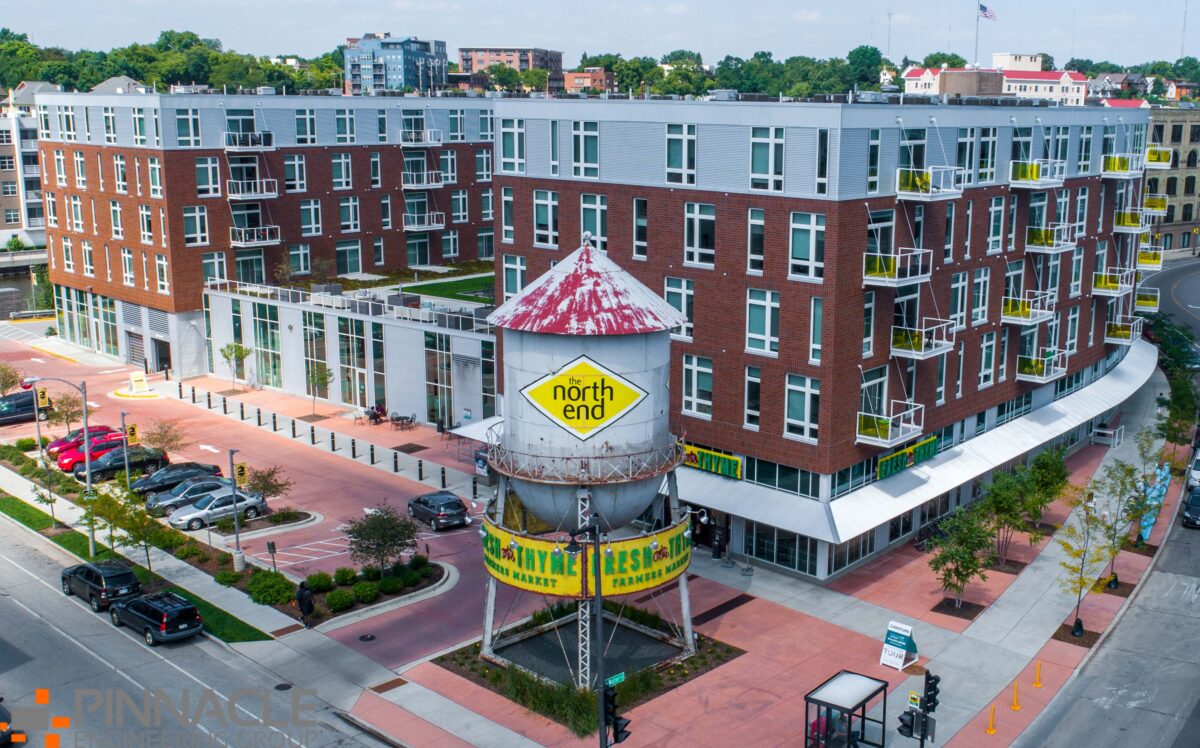 A red-brick apartment building with yellow balcony railings is behind a vintage water tower marked north end. The water tower base advertises a farmers market. The scene includes trees, streets, and nearby parked cars.