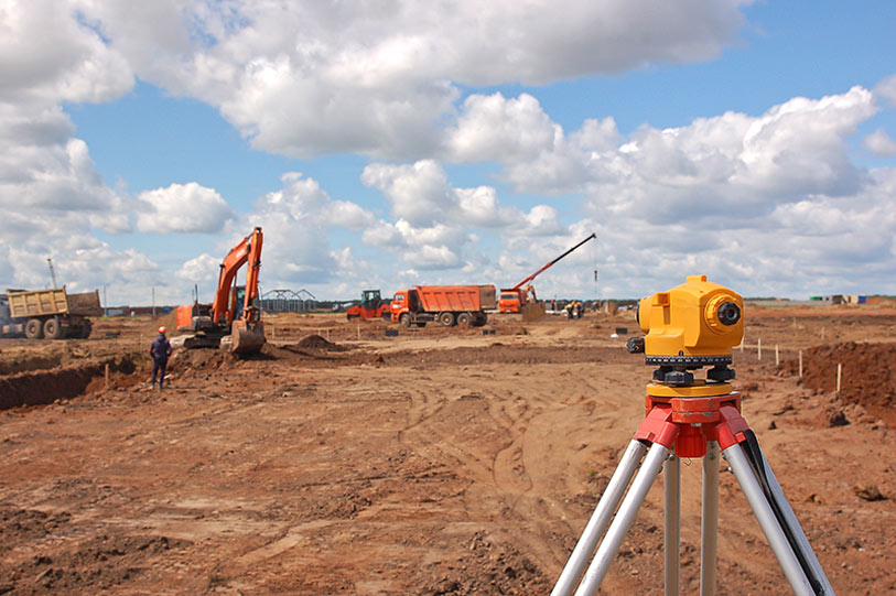 A yellow surveying instrument stands on a tripod in the foreground of a construction site with excavators, dump trucks, and a worker visible under a partly cloudy sky.