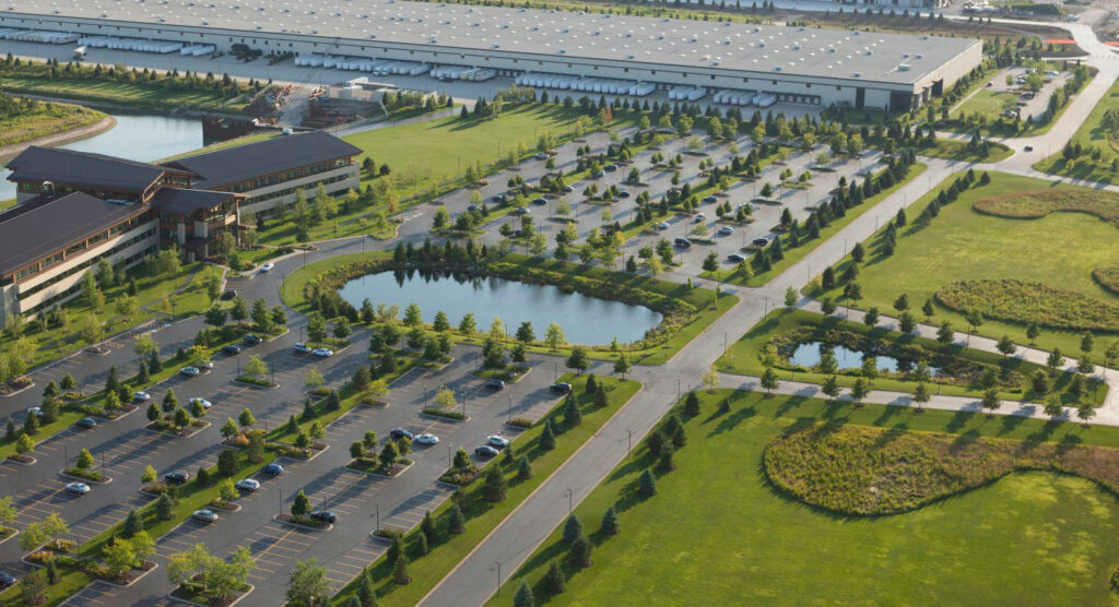 Aerial view of a large office or industrial complex with several parking lots, ponds, landscaped green areas, and a long warehouse building in the background. Few cars are parked, and trees line the roads and lots.