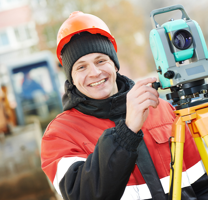 A smiling construction worker in a red jacket and orange hard hat operates a teal surveying instrument outdoors, with blurred construction equipment and buildings in the background.