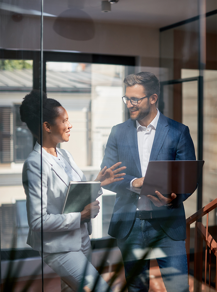 Two business professionals, a woman holding a folder and a man holding a laptop, stand in a modern office by a window, smiling and engaged in conversation. Sunlight streams in, creating a bright, professional atmosphere.