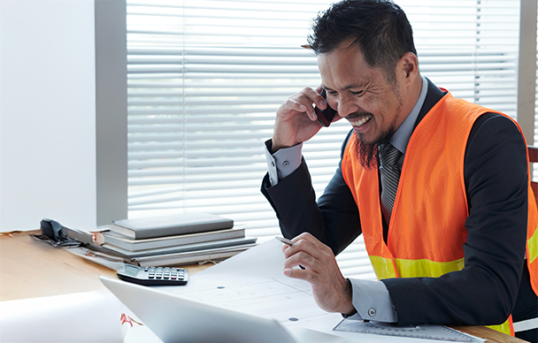 A man in a safety vest smiles while talking on the phone and looking at paperwork at his desk. A calculator, folders, and a laptop are also on the desk, and blinds cover the window behind him.