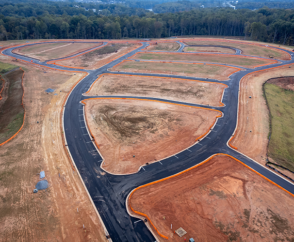 Aerial view of a construction site with newly paved roads and outlined plots of land surrounded by orange barriers, bordered by green trees in the background.